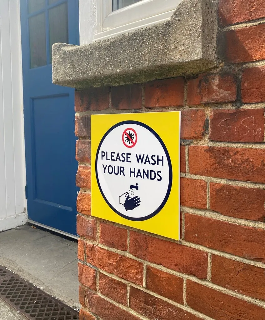 Please Wash Your Hands sign fixed to the red brick wall beside a school entrance