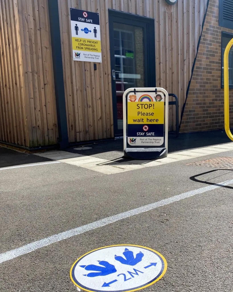 A-board reading STOP Please Wait Here beside a school entrance, with a 2M floor graphic on the path