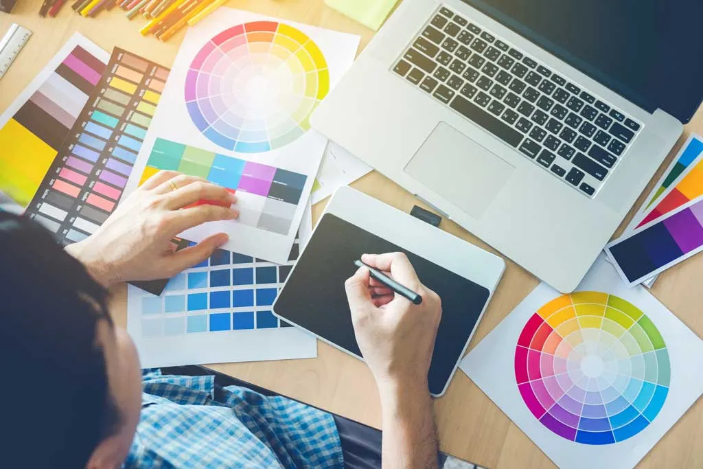 A designer working at a graphics tablet and laptop surrounded by colour wheels and swatch charts