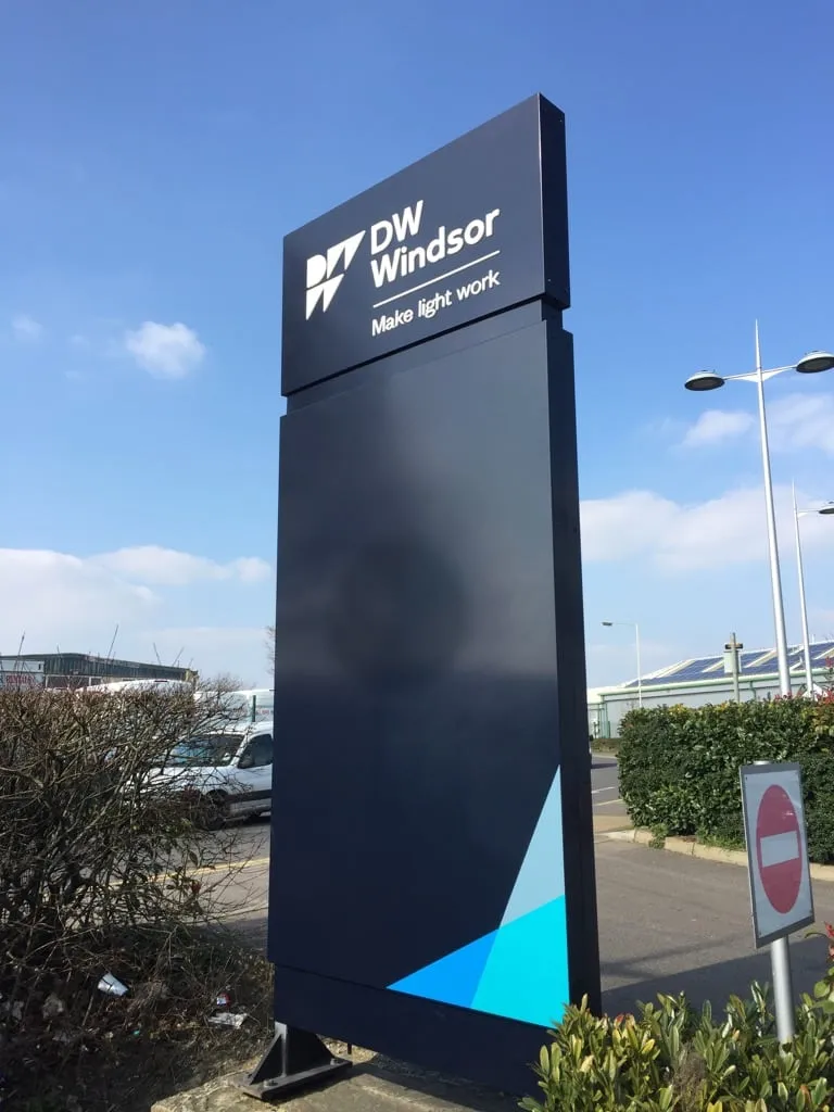 A tall dark totem sign for DW Windsor standing in a car park against a blue sky