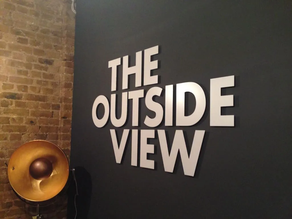 White raised letters reading The Outside View on a dark wall beside a copper wall lamp