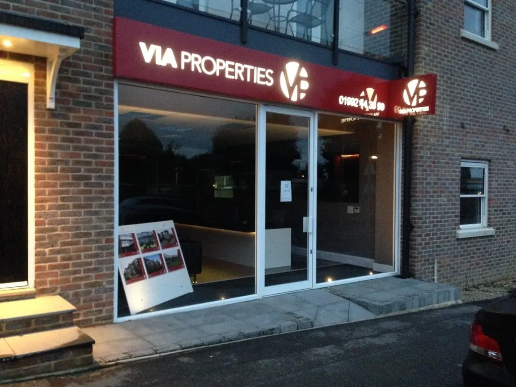 A red illuminated VIA Properties fascia sign above an estate agent's glazed frontage