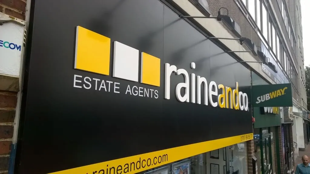 A black estate agent fascia sign carrying the raine and co logo in yellow and white